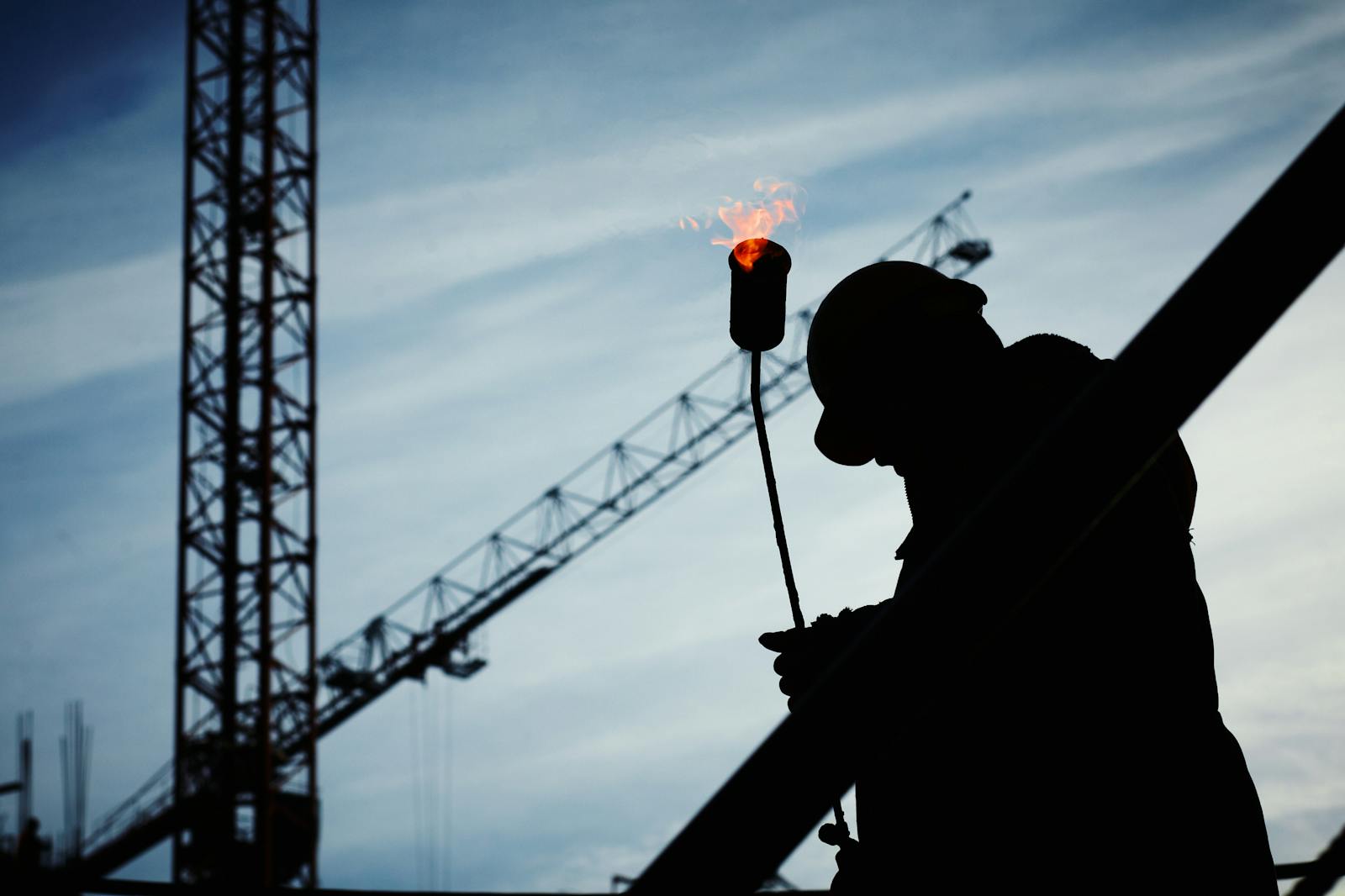 Silhouette of a construction worker using a blowtorch at a building site against a crane-filled skyline. commercial contractors