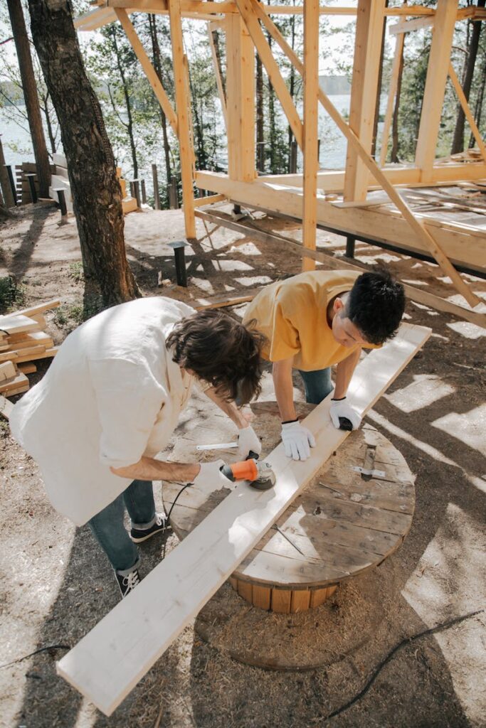 Two adults woodworking outdoors, using power tools on a wooden plank.