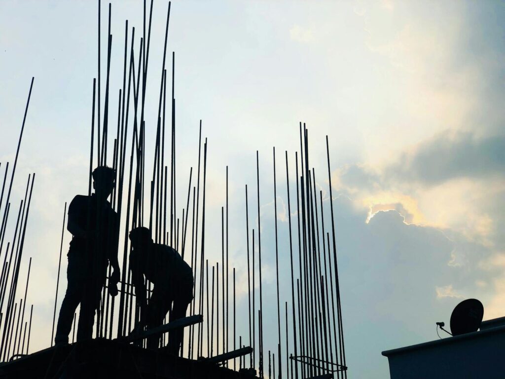 Silhouetted construction workers on site at dawn in Dhaka, Bangladesh, against a cloudy sky.
