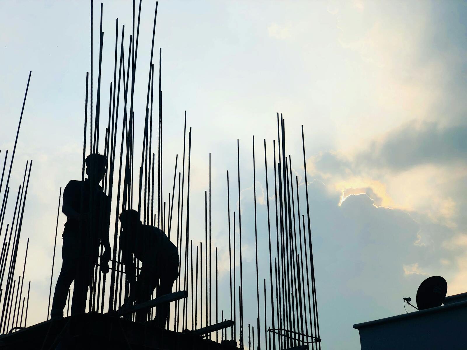 Silhouetted construction workers on site at dawn in Dhaka, Bangladesh, against a cloudy sky.