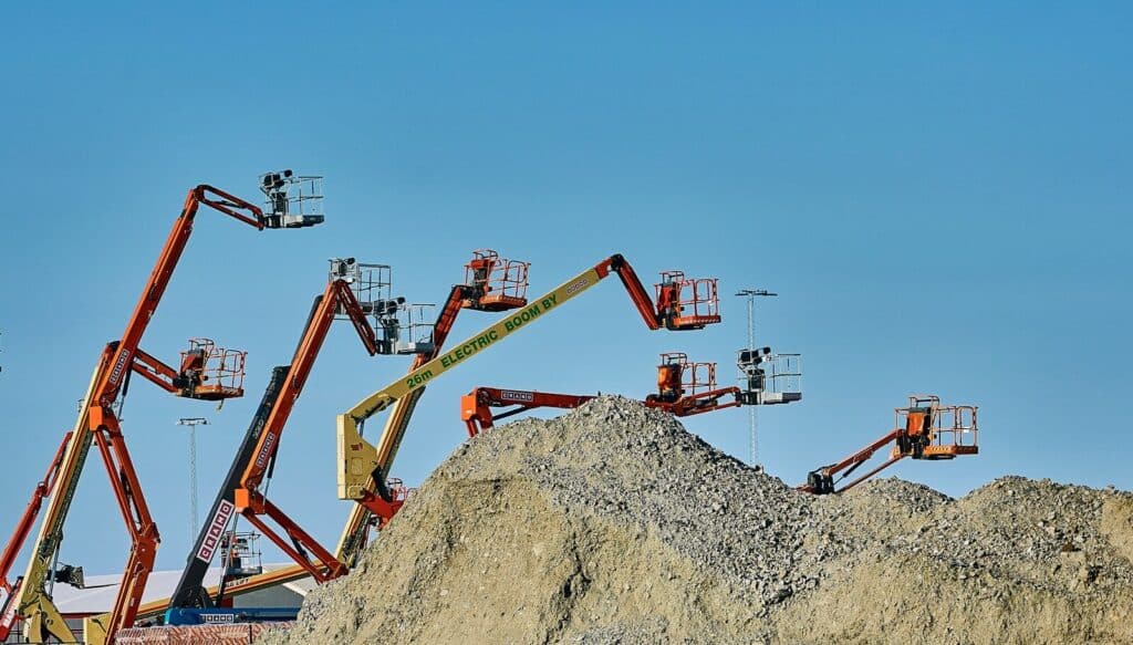 Several orange and yellow boom lifts positioned behind a large pile of gravel.
